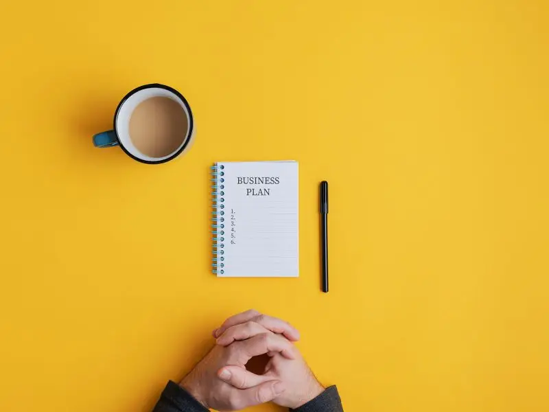 A Time to Review and Plan. This image shows a bright yellow background with a business man sat at the desk with a cuppa a pen and a notepad with the wording Business Plan and 5 blank steps on it.