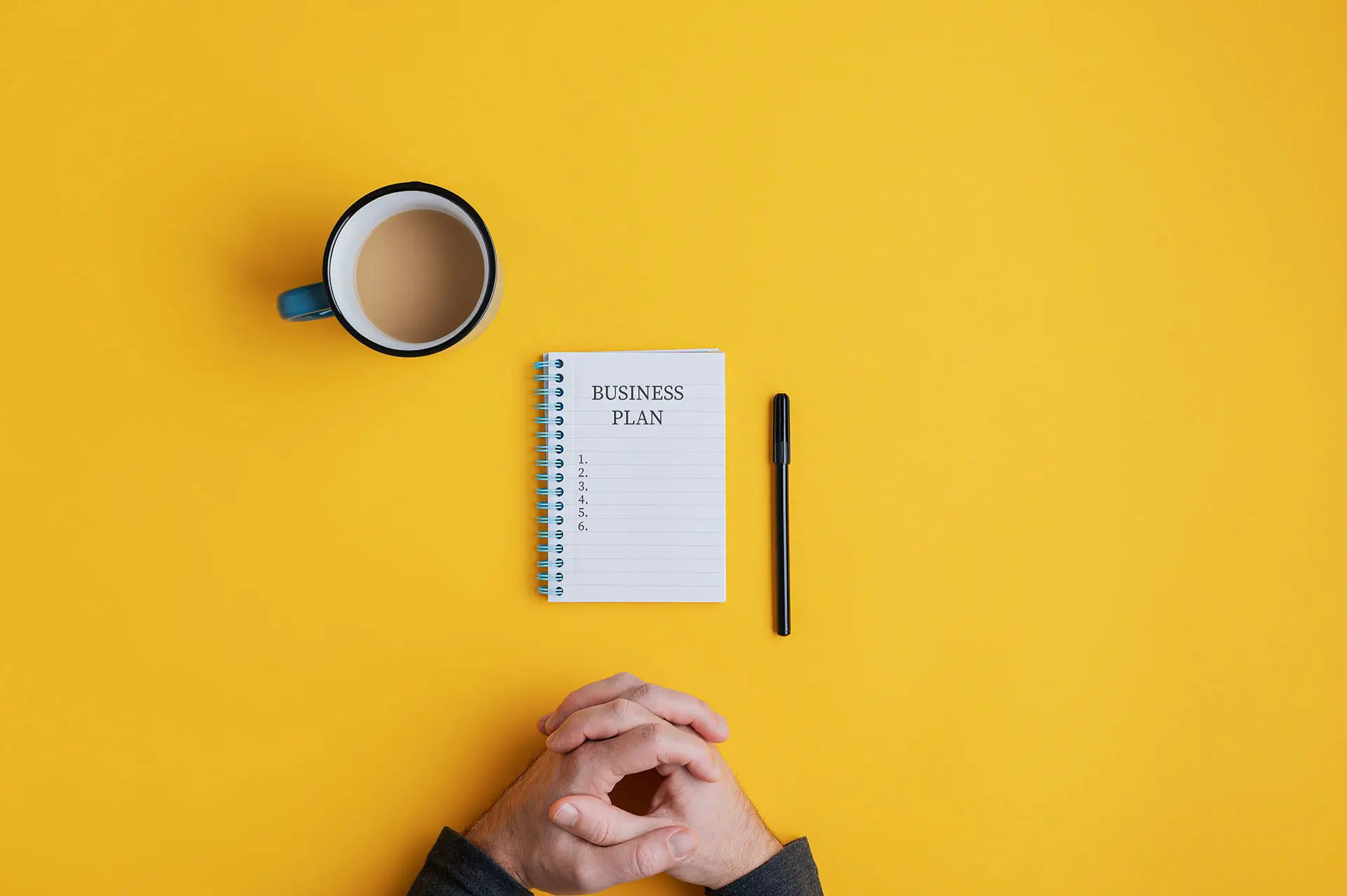 A Time to Review and Plan. This image shows a bright yellow background with a business man sat at the desk with a cuppa a pen and a notepad with the wording Business Plan and 5 blank steps on it.