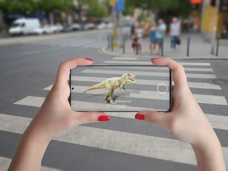 Augmented Reality the image shows hands holkding a smartphone over a zebra crossing the screen shows a dinosaur that isn't there in real life