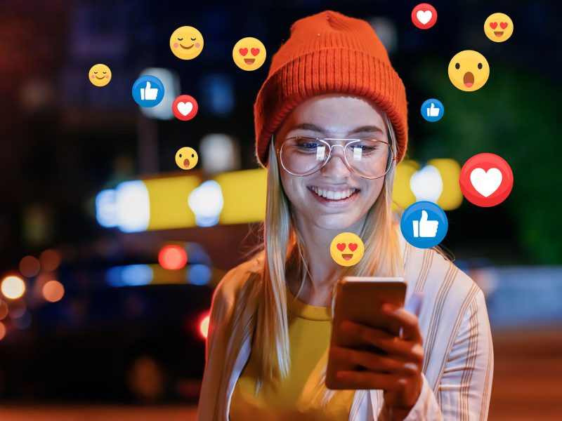 Business Growth Through Facebook - this image shows a lady at night with an ornage hat and glasses, she is looking at her mobile phone and smiling at there are facebook icons around the phone