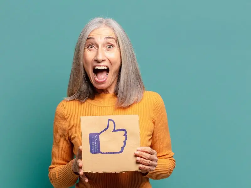 Facebook Save Feature, this image shows a happy, mature lady in a yellow jumper holding a piece of cardboard with a thumbs up on it - Facebook save feature concept