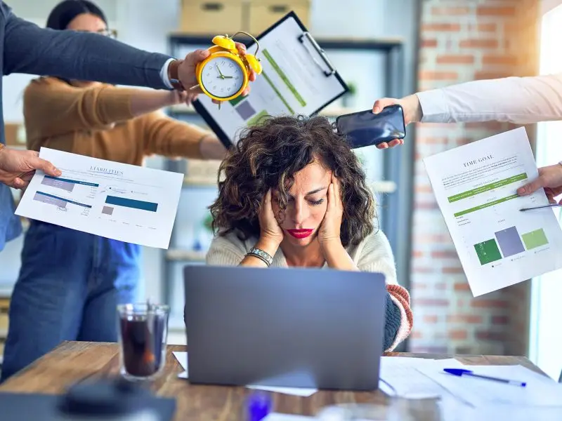 How To Manage Your Time More Effectively. This image shows an overwhelmed lady at her desk with her head in her hands whilst people around her reach out with reports, goals, a clock and other items. Time management concept