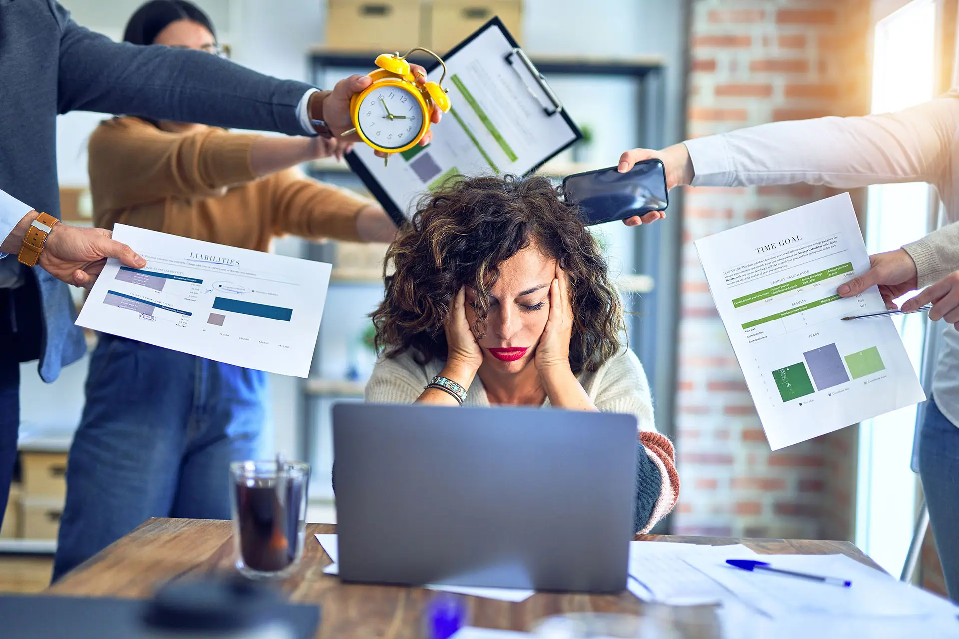 How To Manage Your Time More Effectively. This image shows an overwhelmed lady at her desk with her head in her hands whilst people around her reach out with reports, goals, a clock and other items. Time management concept