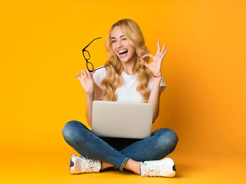 How to Write a Great Case Study, the image shows a lady sat on the floor cross legged with a laptop in her lap, she is holding her glasses with one hand and making an OK sign with her other