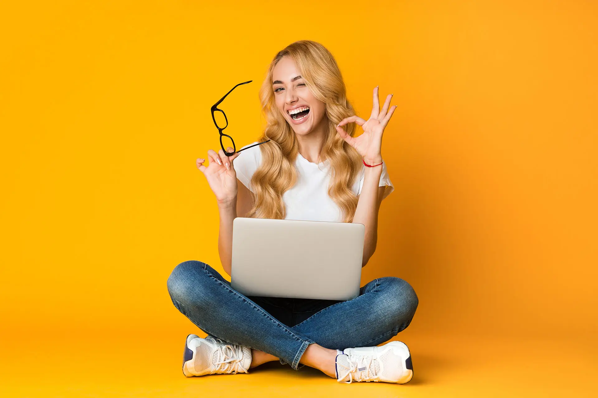 How to Write a Great Case Study, the image shows a lady sat on the floor cross legged with a laptop in her lap, she is holding her glasses with one hand and making an OK sign with her other