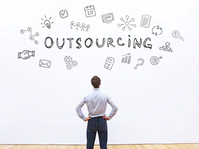 Small Businesses Need to Outsource to Survive! This image shows a business man stood before a white wall which holds the word "outsourcing" and various icons that depict ides, growth, sales, marketing, IT etc.