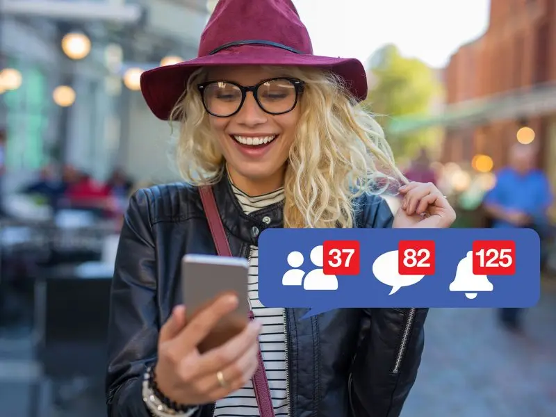 Social Media Numbers this image shows a happy lady on a town high street, she is wearing a plum coloured hat, glasses and is holding her phone in her hand, she is smiling at the content and social media notification icons are displayed with follows, comments and alerts - concept for social media numbers