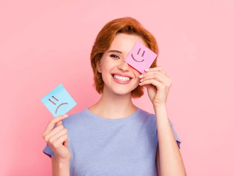 The Importance of Good Customer Communications. This image shows a pink background with a red head lady holding two sticky notes, one with a frown face drawn on it and the other with a happy face, she is smiling and holding the happy sticky note up to her face.