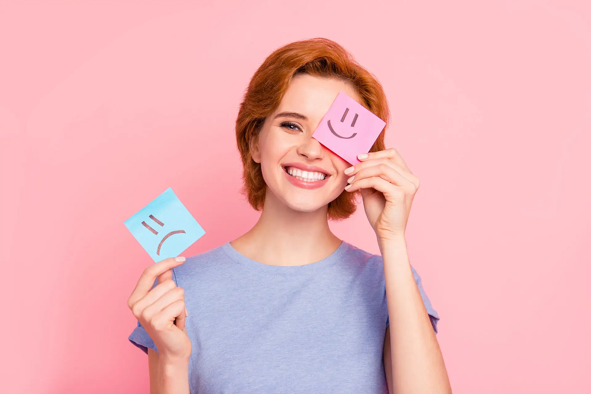 The Importance of Good Customer Communications. This image shows a pink background with a red head lady holding two sticky notes, one with a frown face drawn on it and the other with a happy face, she is smiling and holding the happy sticky note up to her face.