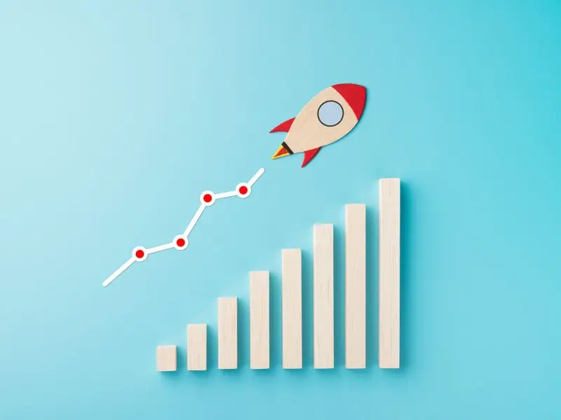 The Other Side of Sales the image shows a pale blue background with a wooden rocket tacking off diagonally climbing a wooden bar chart - sales concept