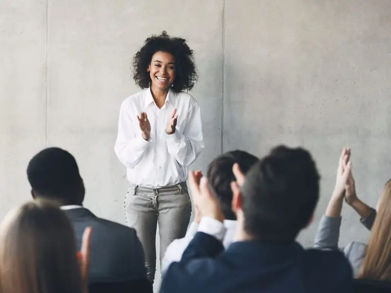 Tips for an Effective Networking Pitch. This image shows a lady stood up speaking at a business event, she is smiling and others are clapping. Concept for Tips for an Effective Networking Pitch