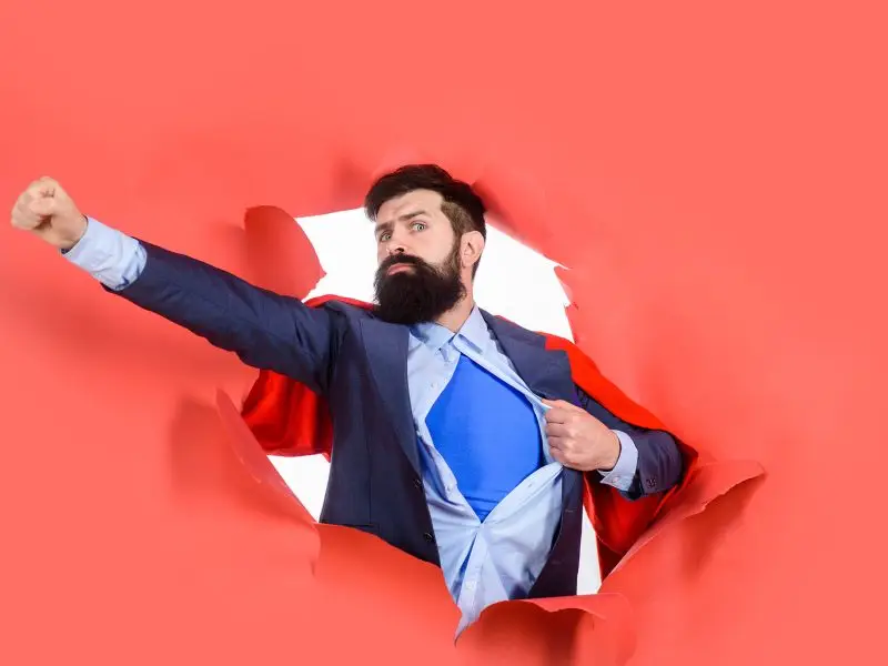 Wearing Underpants on the Outside the image shows a bearded man with a red cape and a blue superhero suit under a business suit, bursting out of a red paper background. Concept for superhero business owner