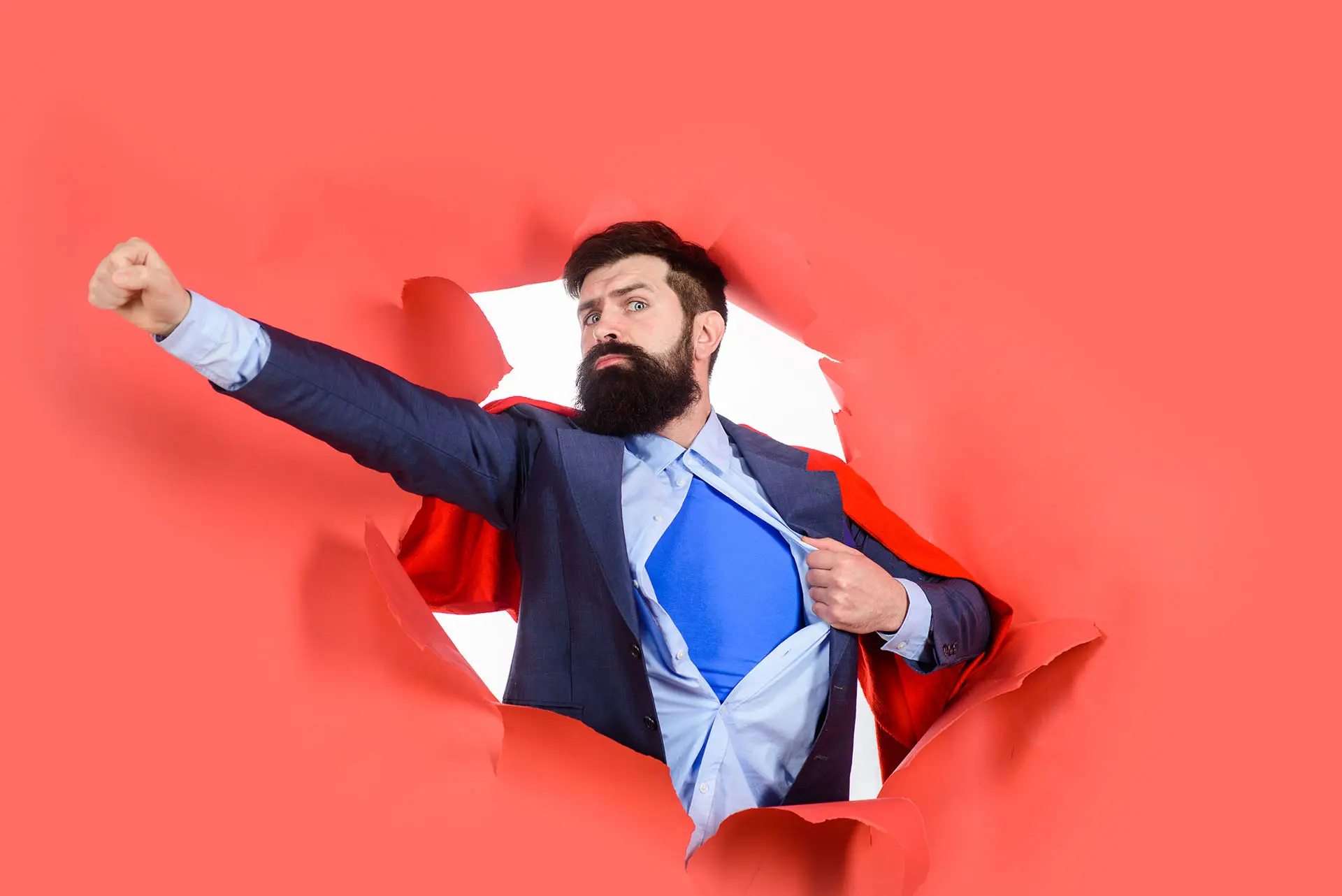 Wearing Underpants on the Outside the image shows a bearded man with a red cape and a blue superhero suit under a business suit, bursting out of a red paper background. Concept for superhero business owner