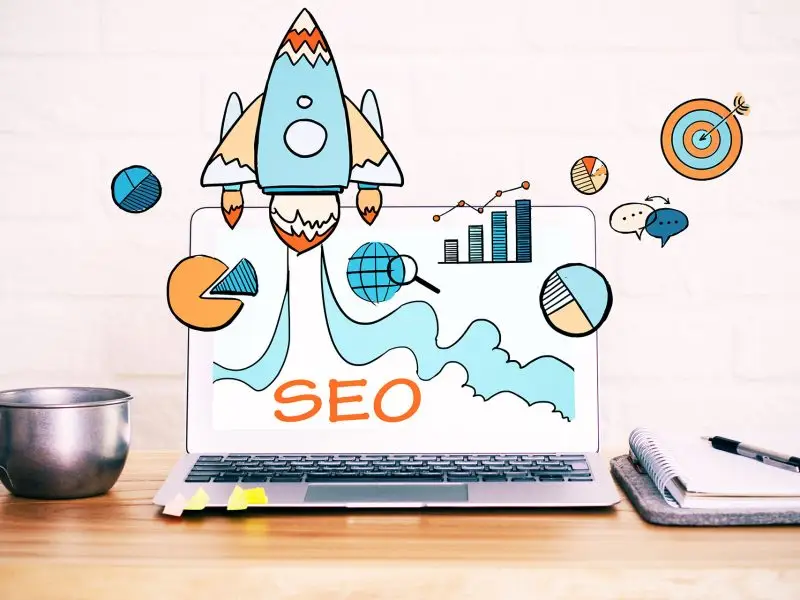 What is a Keyword Research? This image shows a laptop on a desk with a rocket graphic and SEO icons and words overlayed on top - keyword concept