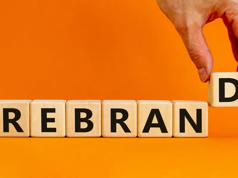 What to Consider Before You Rebrand - this image shows a vibrant orange background with the word rebrand spelled out in blocks, with a hand lifting in the last letter