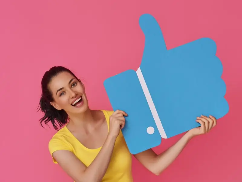 A Guide to Using Facebook Ads - this image shows a btight pink background with a lady in a bright yellow top holding and over-sized blue thumb - concept for Facebook ads