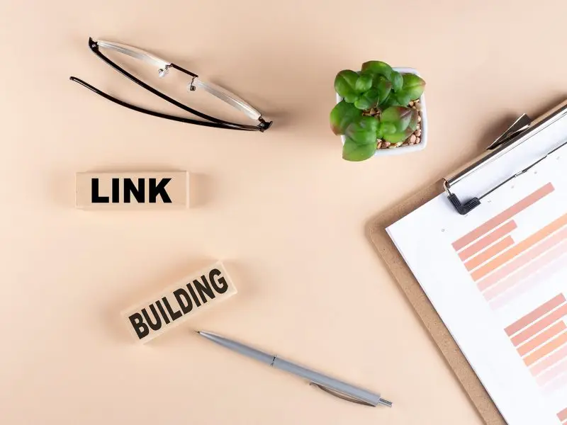 Are Backlinks Good or Bad for SEO? This image shows a peach desk with glasses, a clipboard, pen and wooden block that say Link Building.