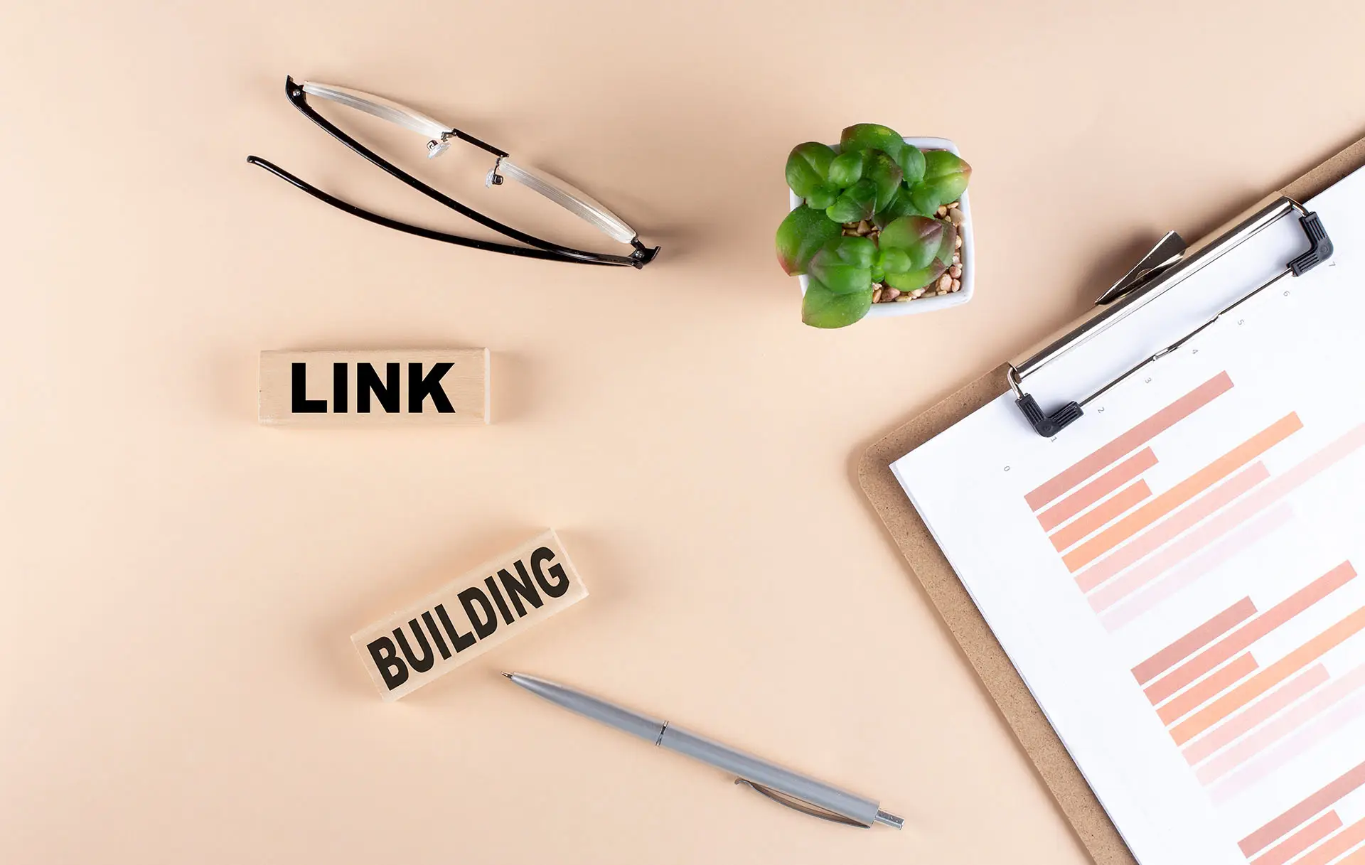 Are Backlinks Good or Bad for SEO? This image shows a peach desk with glasses, a clipboard, pen and wooden block that say Link Building.