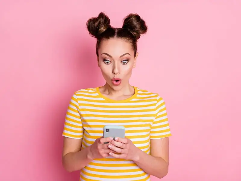 Facebook Ads - How to Create Powerful Copy. This image shows a bright pink background with a lady in a yellow and white striped Tshirt looking at her mobile, her expression is wow - concept for powerful ad content