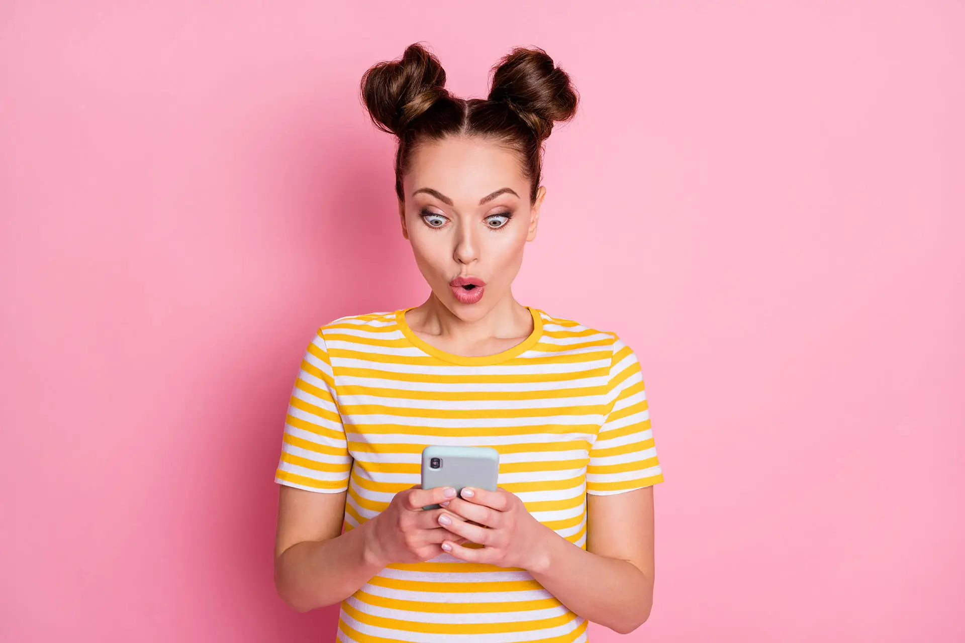 Facebook Ads - How to Create Powerful Copy. This image shows a bright pink background with a lady in a yellow and white striped Tshirt looking at her mobile, her expression is wow - concept for powerful ad content