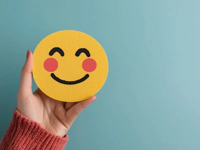 SEO Versus User Engagement - this image shows a light blue background with a hand holding a yellow smiling emoji - concept for balancing user experience and seo