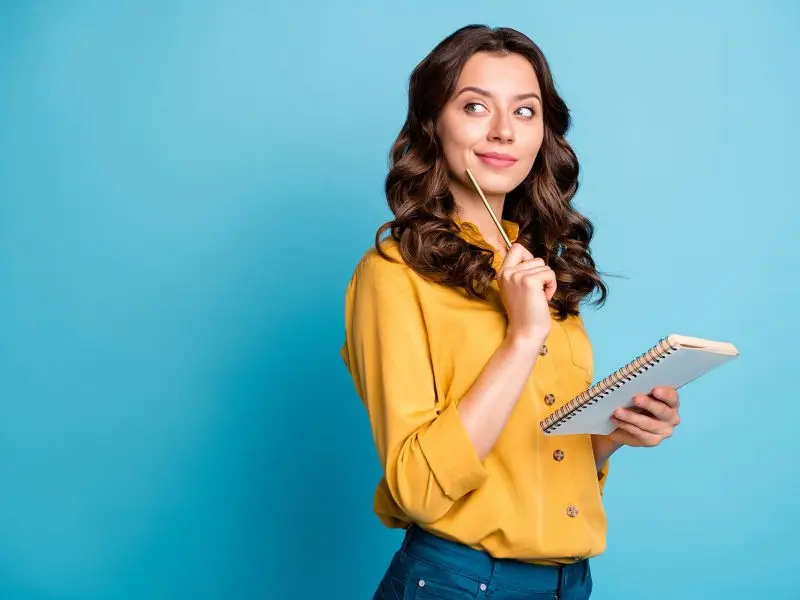 The Benefits Of Having Your Blog On Your Website. This image shows a bright blue background, with a lady in a yellow shirt in the foreground, she is holding a notpad and a pencil and has raised the pencil up to indicate a good idea or thought process