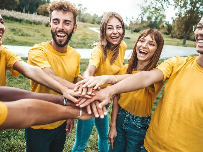 The Business Benefits of a Social Conscience. This image shows a team of people all in yellow shirts volunteering, they all have their hands in a huddle and are all smiling. Volunteering concept