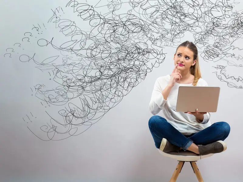 Website Jargon Explained. This image shows a lady sat cross legged on a chair with a laptop, she looks puzzled and behind her are various scribble lines ending in question marks - concept for Website Jargon Explained