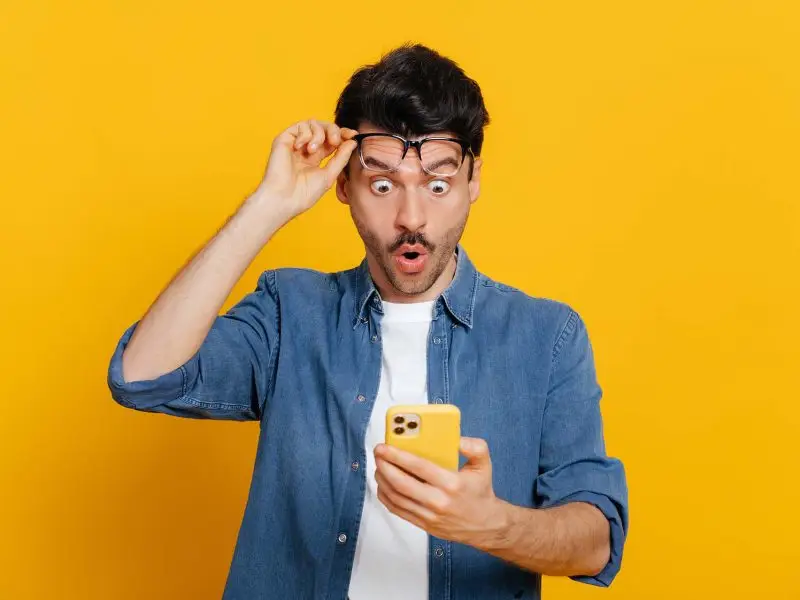 Creating Landing Pages that Convert - this image shows a bright yellow background, with a man in a denim shirt in the foreground, he is lifting his glasses up to see his mobile phone