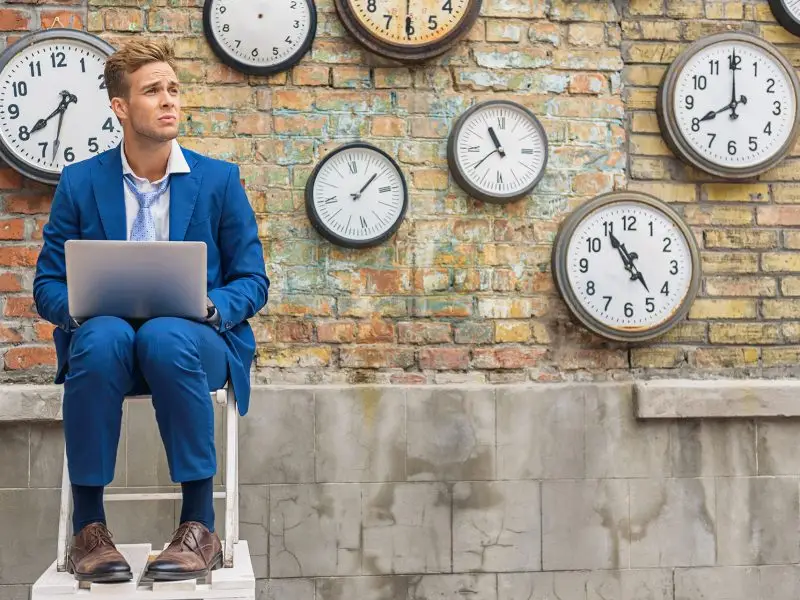 Google Ads Timing is Everything - this image shows a man sat on a wall with a laptop in his lap, behind him on a brick wall are various clocks set to different times - concept for Google Ad timings