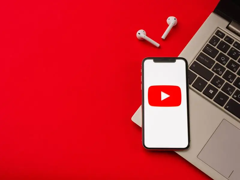 How to Remove the Suggested Video Features in YouTube - this image shows a bright red background with a laptop, ear buds and a mobile with the YouTube player logo