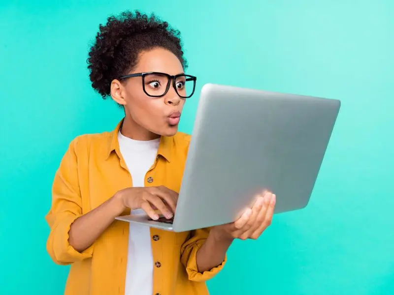 How to Write a Press Release - this image shows a bright blue background with a lady in a bright yellow shirt, holding and using her laptop, her face has an engaged expression