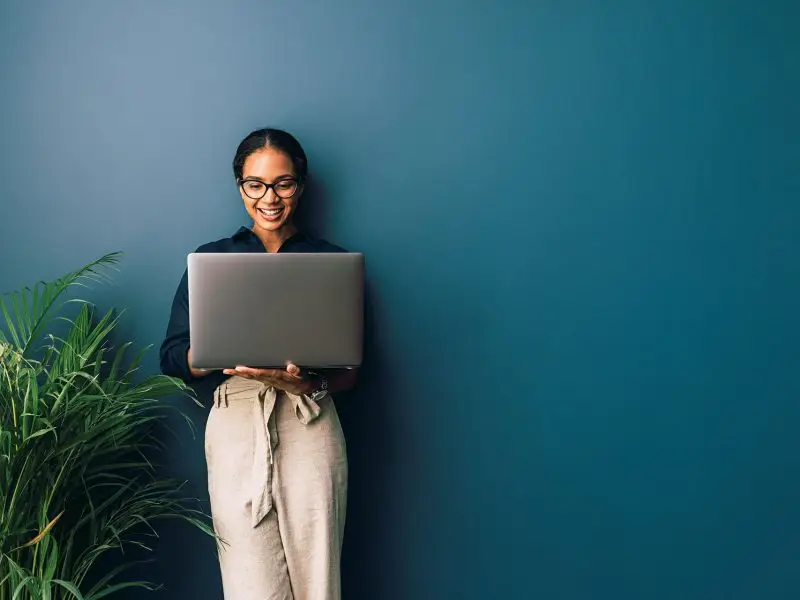 Introducing Workplace by Facebook - this image shows a lady leaning against a petrol blue wall holding her laptop and smiling