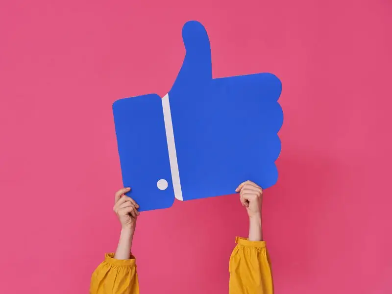 Is Facebook for Business a Waste of Time? This image shows a bright pink background with a lady's arms raised holding a blue Thumb's up logo just like the Facebook icon