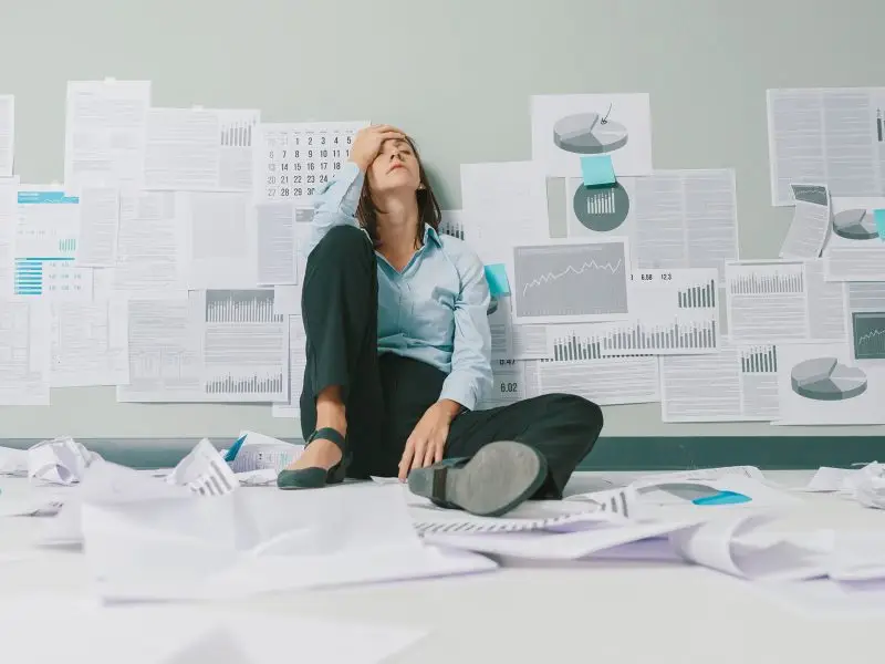 Know the Meaning Behind Website Data - this image shows a woman sitting on the floor with her back against the wasll, she has a knee raised and is resting her arm on it with her head in her hand. There are data graphs pinned to the wall and crumpled up all over the floor.