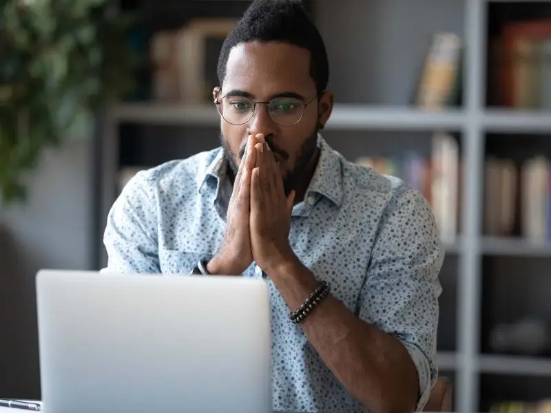 Protecting Your WordPress Website - this image shows a man in a smart shirt sat at a desk with his laptop in fron of him, he has both hands raised to his face and he looks worried - concept for protecting your website