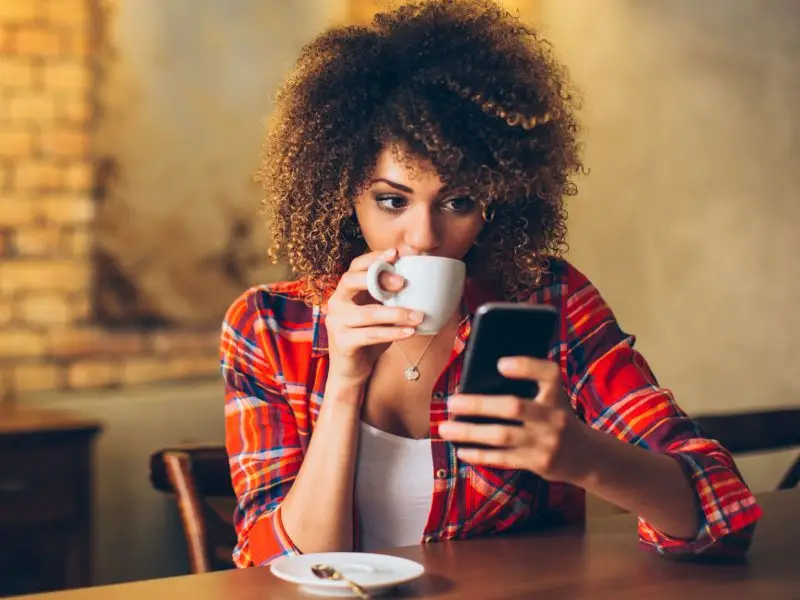 Social Media UK User Statistics for 2020 - this image shows a young lady sat a t a table drinking hot beverage whilst scrolling on her phone