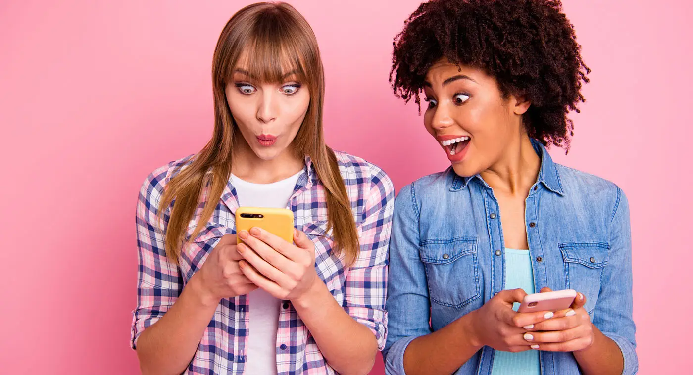 Social Media UK User Statistics for 2020 - this image shows a bright pink background with two ladies using mobile phones, one is looking at the mobile making an "ooo" expression and the other one is looking over her shoulder smiling.