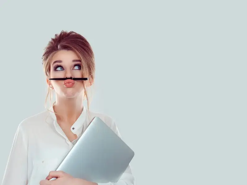 Tell Your Story - this image shows a light grey background, with an attractive young lady holding a laptop and a pen which is being held by her upper lip, this image is a light hearted concept for story telling