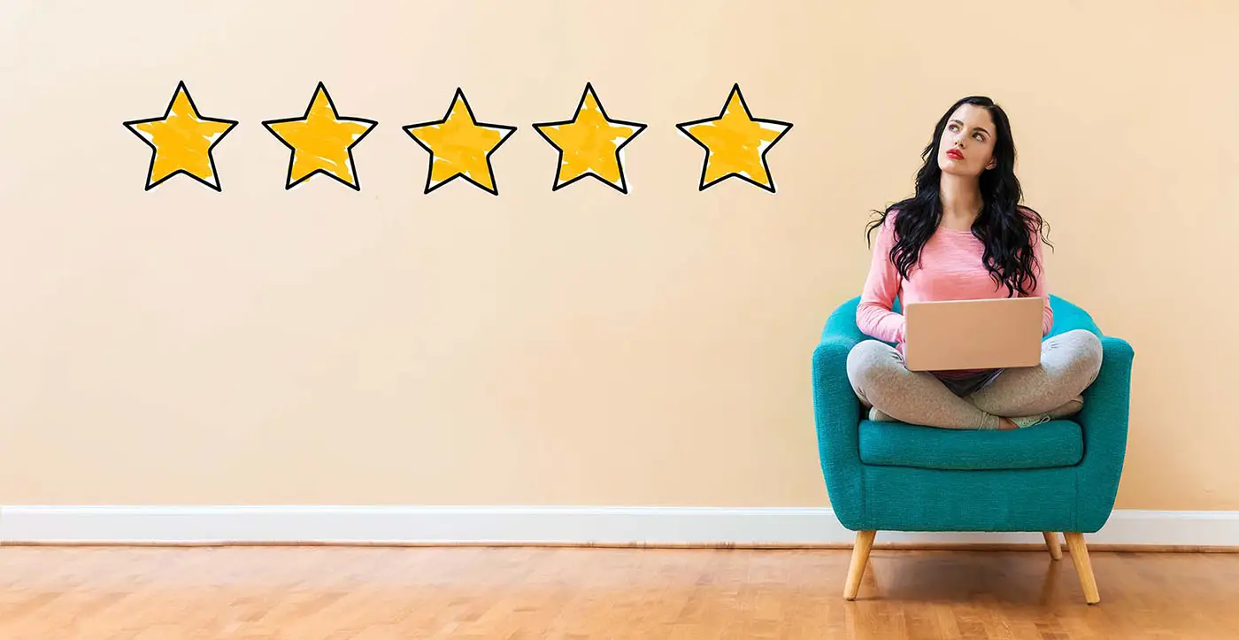 The Importance of Customer Reviews and How to Encourage Them - this image shows a pale yellow background with a lady sat cross legged in an arm chair, she has a laptop in her lap and a thoughtful expression, next to her are 5 gold stars