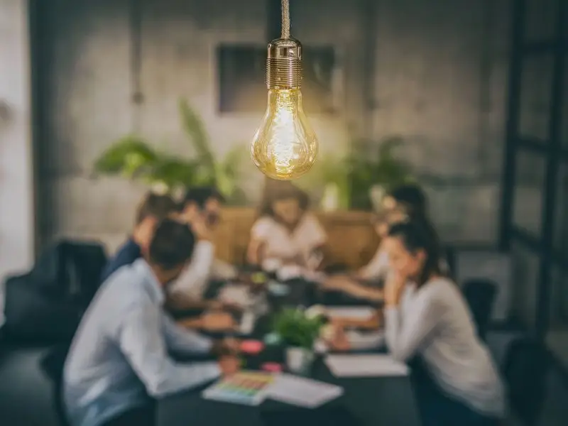 The Social Element of Social Media - this image shows a twam sat chatting around a board table with a lit light light bulb in the foreground, concept for being sociable on social media