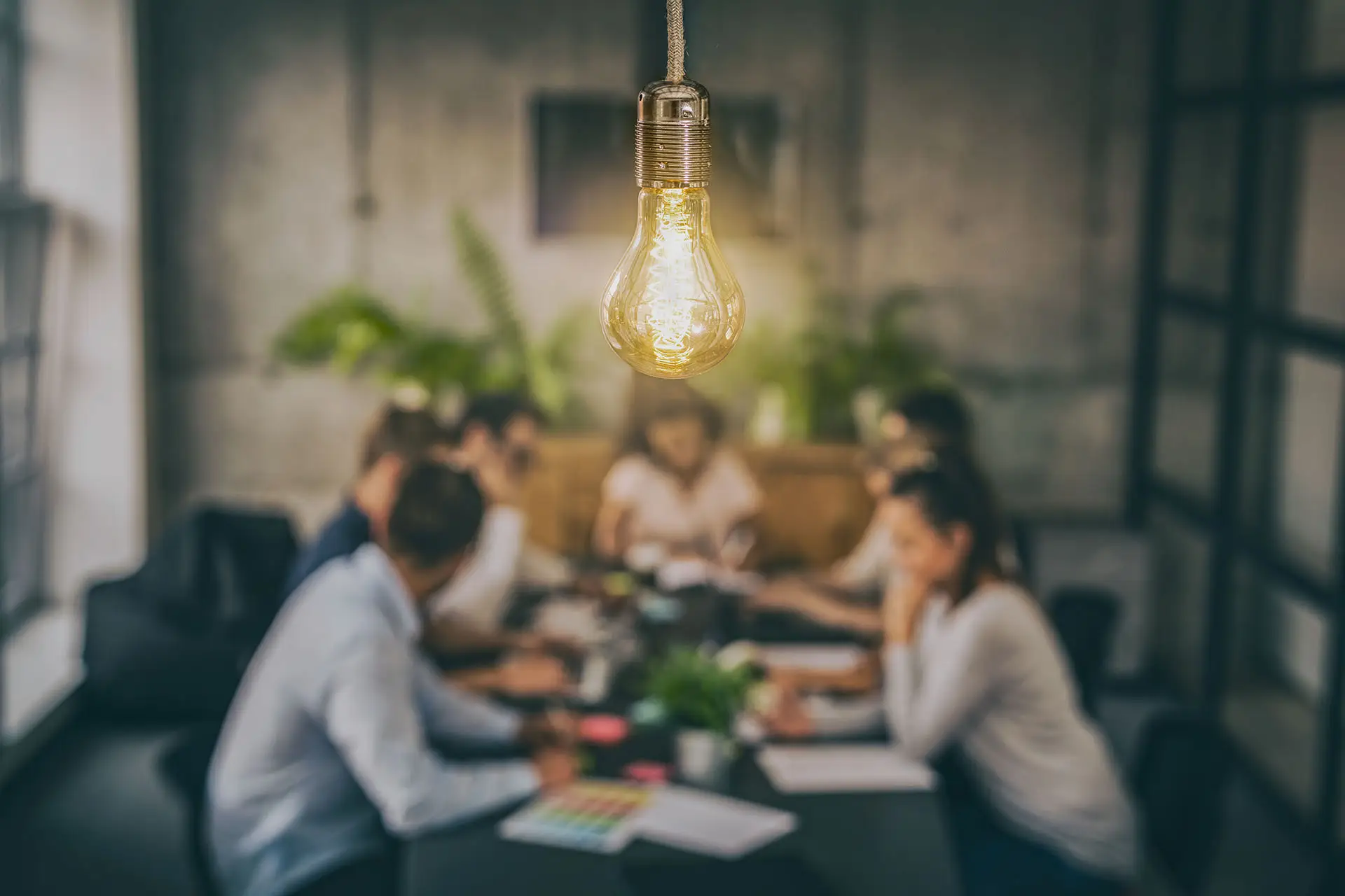 The Social Element of Social Media - this image shows a twam sat chatting around a board table with a lit light light bulb in the foreground, concept for being sociable on social media