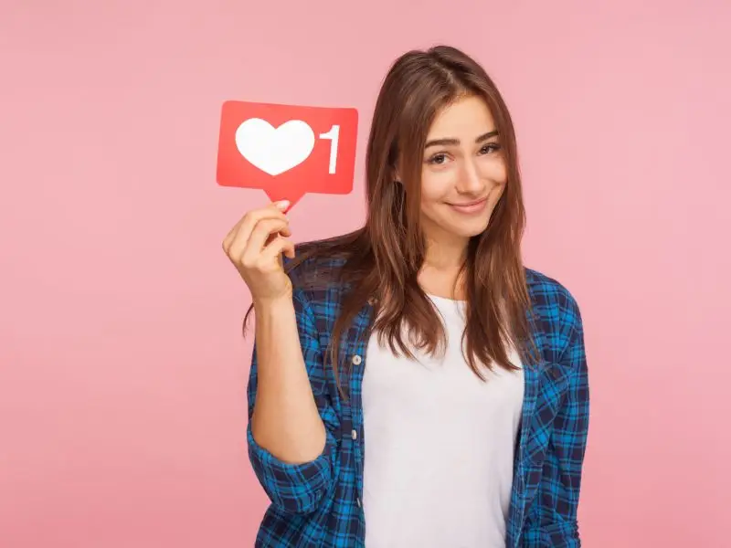 Tips for Growing Your Social Media Accounts - this image shows a bright pink background with a lady in a checked shirt smiling and holding up a social media heart icon