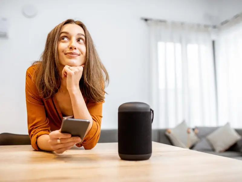 Voice Search Optimisation - this image shows a living room with a lady leaning on a table with her mobile in her hand, on the table is a voice assistant device.