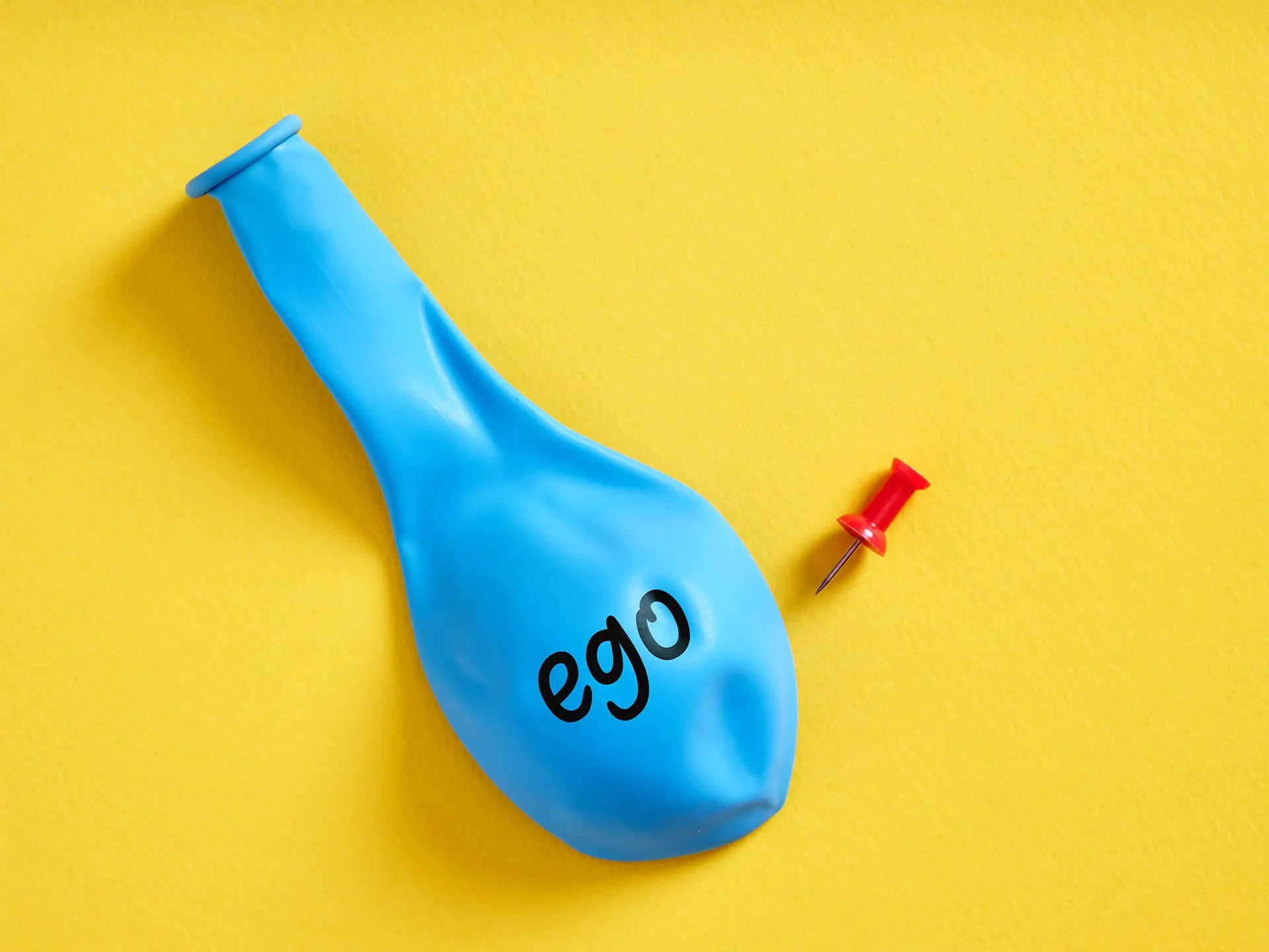 Why Giving Clients What They Want Isn't Always the Best Strategy - this image shows a bright yellow background with a deflated blue baloon with the word ego written on it and a red pin next to it