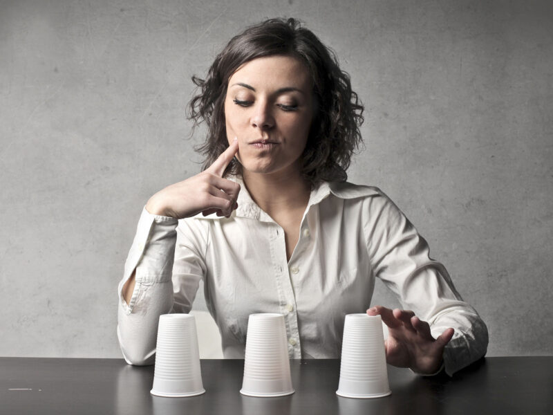 Marketing Without Guesswork - this image shows a grey background, with a lady in a white blouse sitting at a desk, in front of her are three cups. She has her hand on one of the cups and the other raised up to her face. She has a thoughtful expression and is trying to guess which cup holds the item