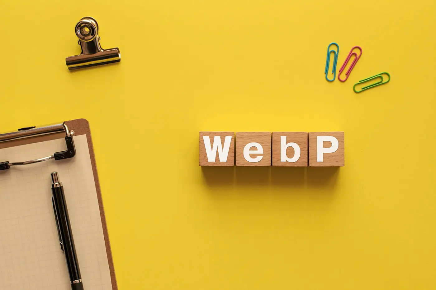 Understanding the Difference Between Lossy and Lossless Compression - this image shows a bright yellow background with a blank clipboard and pencil some paperclips and wooden blocks that spell out WebP