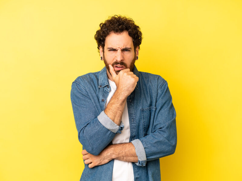 What Are Landing Pages? This image shows a bright yellow background with a confused man in the foregraound with one arm across his body and the other raised with his hand cupping his chin, he has a thoughtful expression on his face