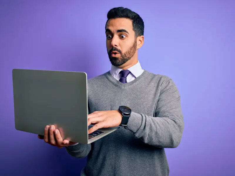 What Should Be Included in an Email Campaign? This image shows a bright purple background with a man in a shirt, tie and grey jumper holding a lamptop, he is looking at the screen with a surprised expression