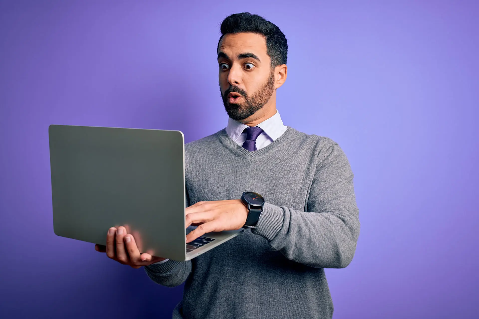 What Should Be Included in an Email Campaign? This image shows a bright purple background with a man in a shirt, tie and grey jumper holding a lamptop, he is looking at the screen with a surprised expression